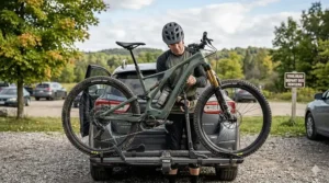 A photorealistic illustration of a rider loading a green full suspension e-bike onto a car rack in an Ontario park, featuring a trailhead sign with bilingual text: "TRAILHEAD / DÉPART DES SENTIERS."