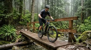 Alt text for image 5: A rider standing with a lightweight hardtail electric mountain bike on a rugged coastal trail in Nova Scotia, with the rocky Atlantic shoreline and crashing waves providing a dramatic backdrop.