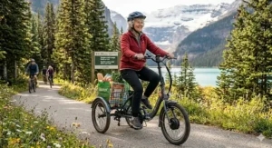 A senior woman riding a foldable electric trike on a dirt trail beside a turquoise glacier lake with snow-capped mountains in the Canadian Rockies.